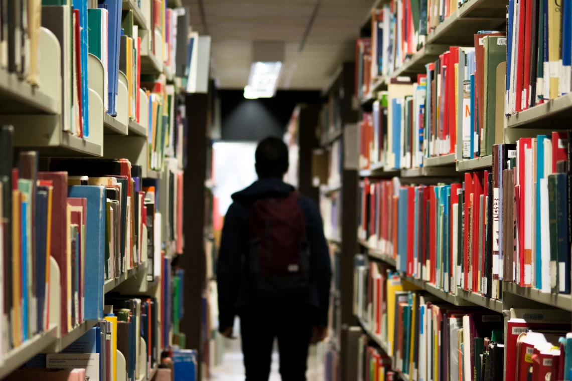 A young man walking through two tall aisles of books.